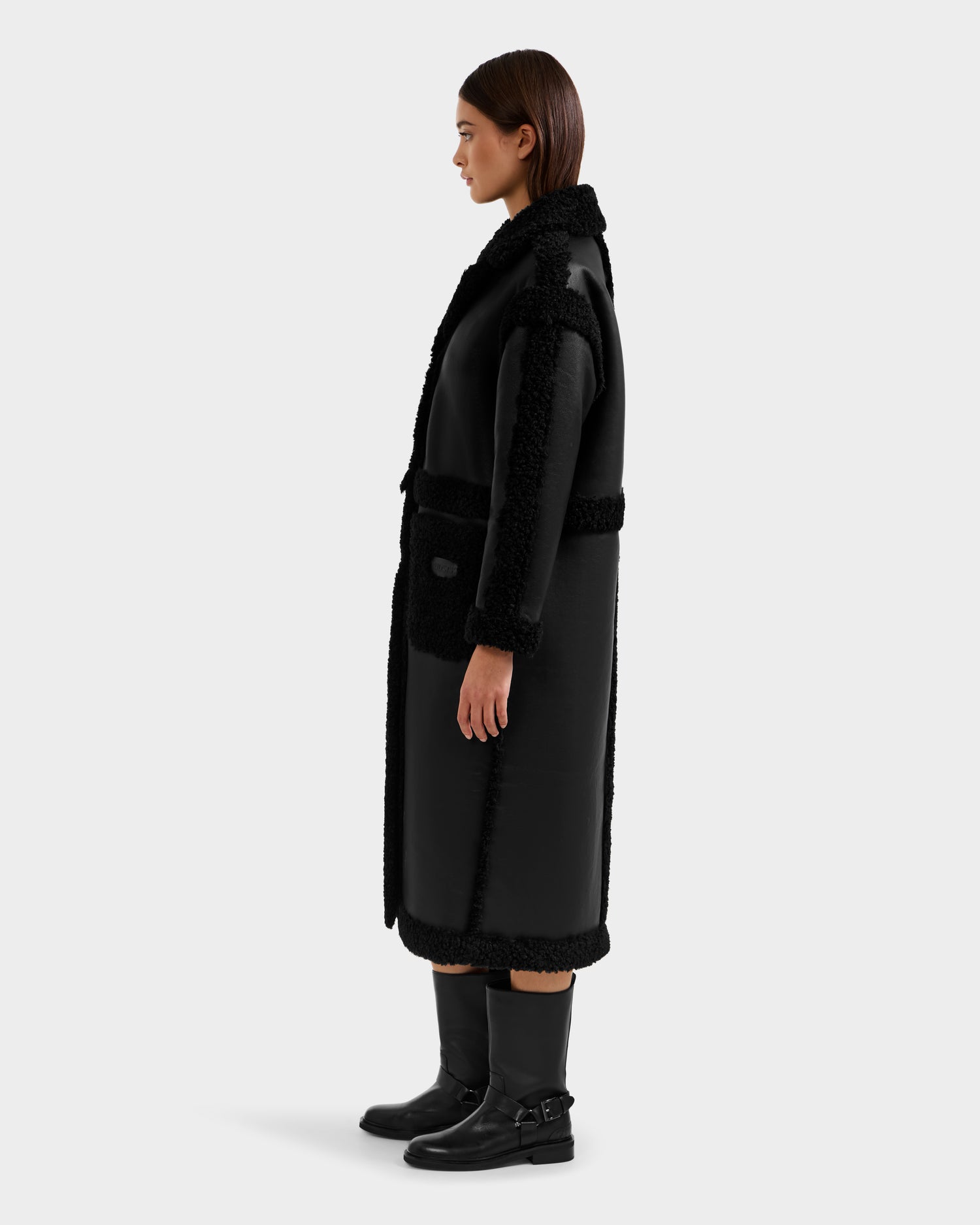 A woman in profile wears the Rudsak PALOMA - 8125675 Black faux shearling coat with textured trim, paired with black mid-calf buckle boots. She has straight brown hair and stands against a plain light gray background.