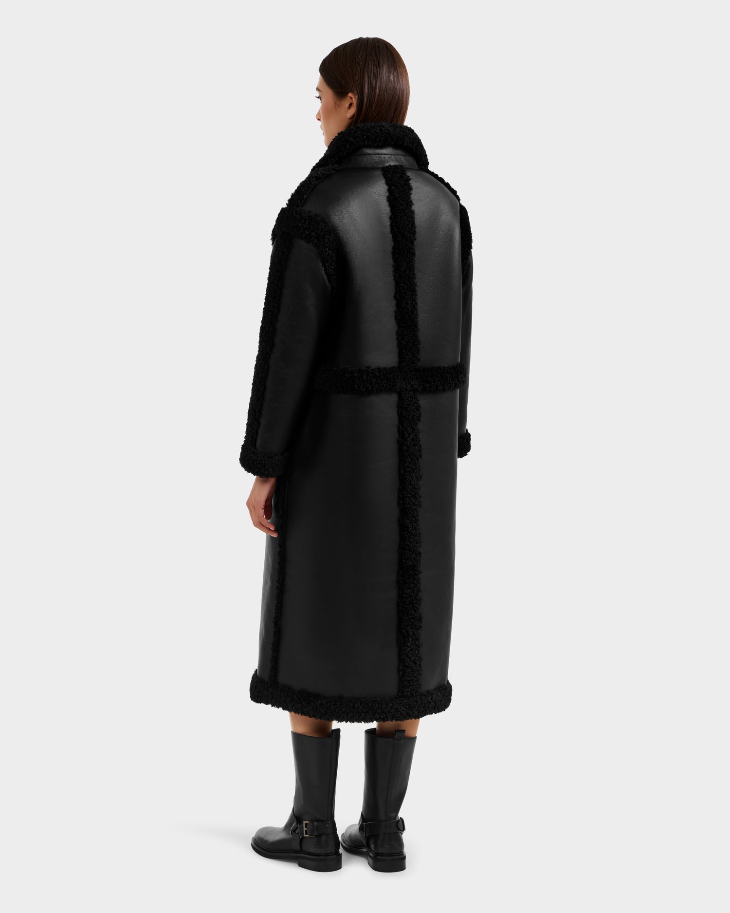 A woman faces away wearing the Rudsak PALOMA - 8125675 Black faux shearling coat with textured trim, wide collar, and straight silhouette. She pairs it with black mid-calf boots against a plain light background.