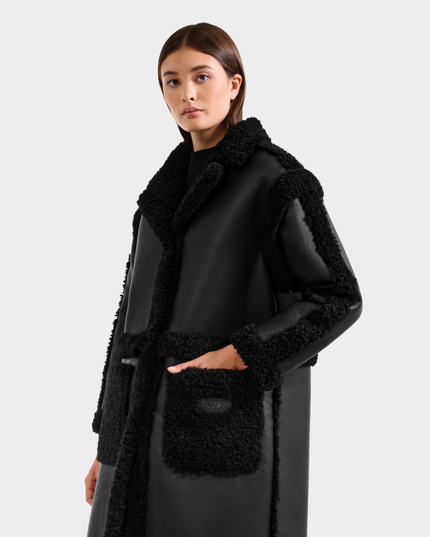 A woman stands confidently against a plain light background, wearing the Rudsak PALOMA - 8125675 Black faux shearling coat with textured trim and large front pockets.
