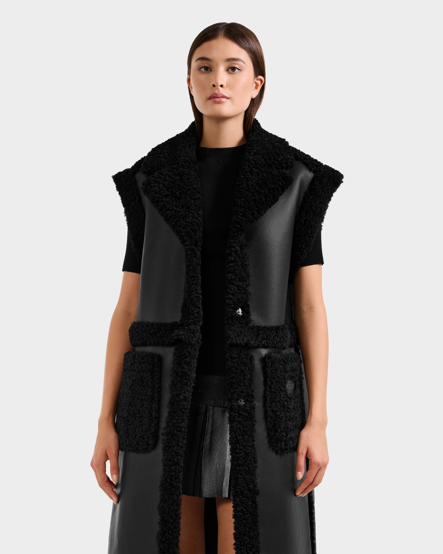 A woman wears the Rudsak PALOMA - 8125675 Black sleeveless faux shearling vest with a large collar and pockets over a black top and skirt, standing against a plain light background. This modular piece adds texture and edge to her look.