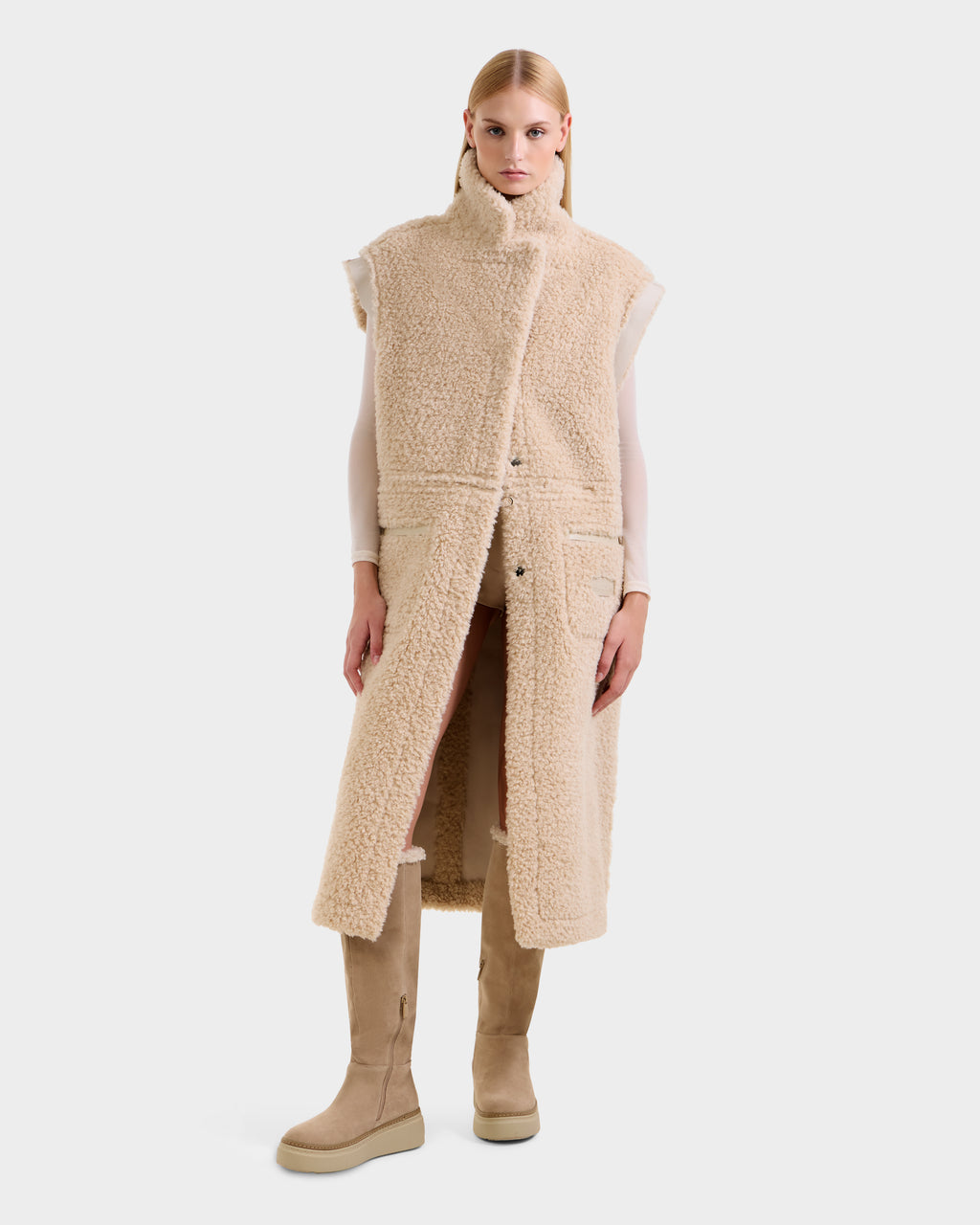 A woman faces forward against a white background, wearing Rudsak's PALOMA - 8125675 Vanilla sleeveless long faux shearling vest over a light top, paired with matching beige knee-high boots. Her hair is straight and sleek.