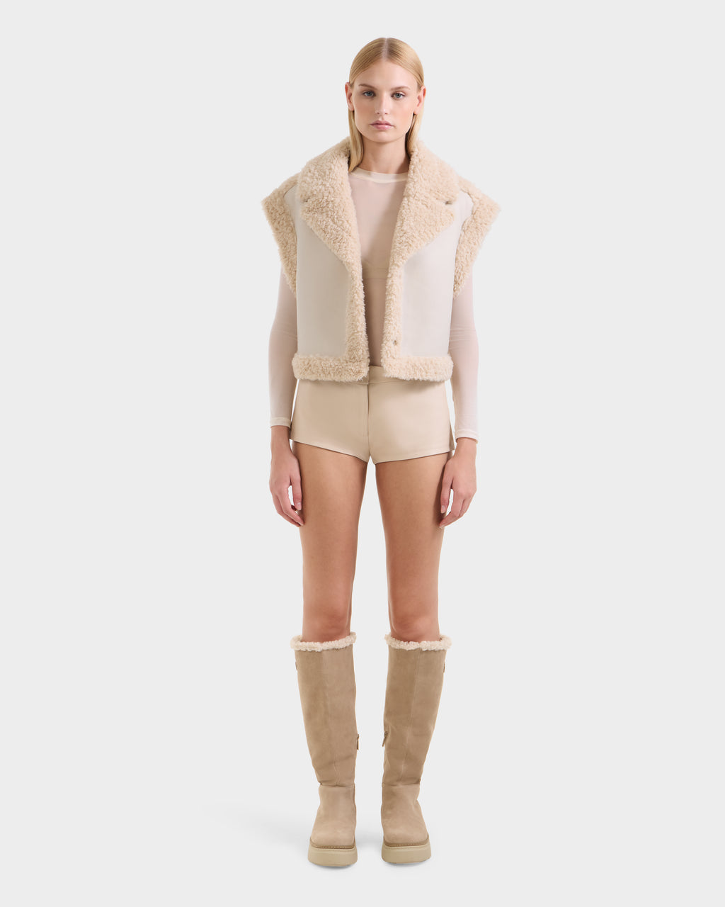 A woman faces forward against a white background wearing the Rudsak PALOMA - 8125675 Vanilla beige faux shearling vest, a sheer long-sleeve top, beige shorts, and knee-high tan boots with shearling trim.