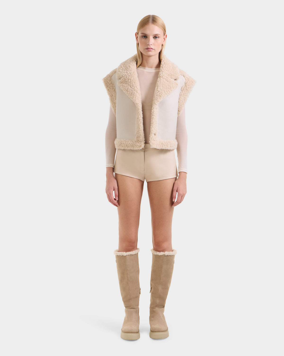 A woman faces forward against a white background wearing the Rudsak PALOMA - 8125675 Vanilla beige faux shearling vest, a sheer long-sleeve top, beige shorts, and knee-high tan boots with shearling trim.