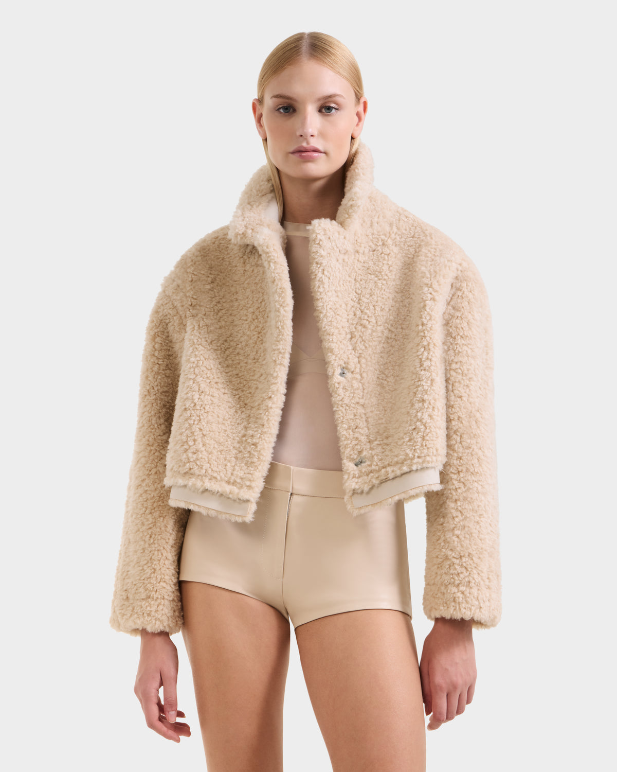 A person wears the Rudsak PALOMA - 8125675 Vanilla faux shearling cropped jacket over a sheer beige top and high-waisted beige shorts, standing facing forward against a plain white background.