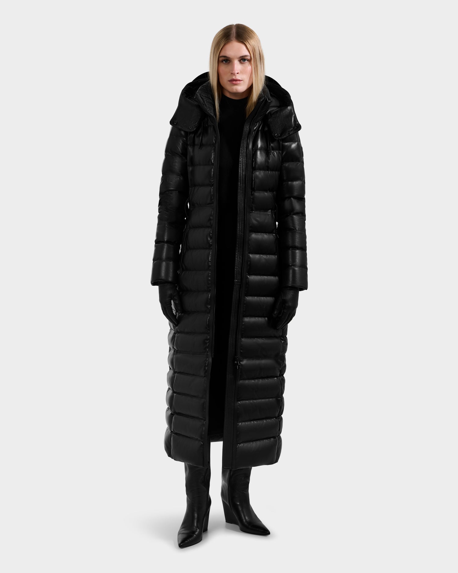 A woman faces forward wearing the Rudsak PELIN LX - 8125692 Black, a shiny black leather down maxi puffer coat with hood, styled with black gloves, dress, and heeled boots against a plain light gray background.