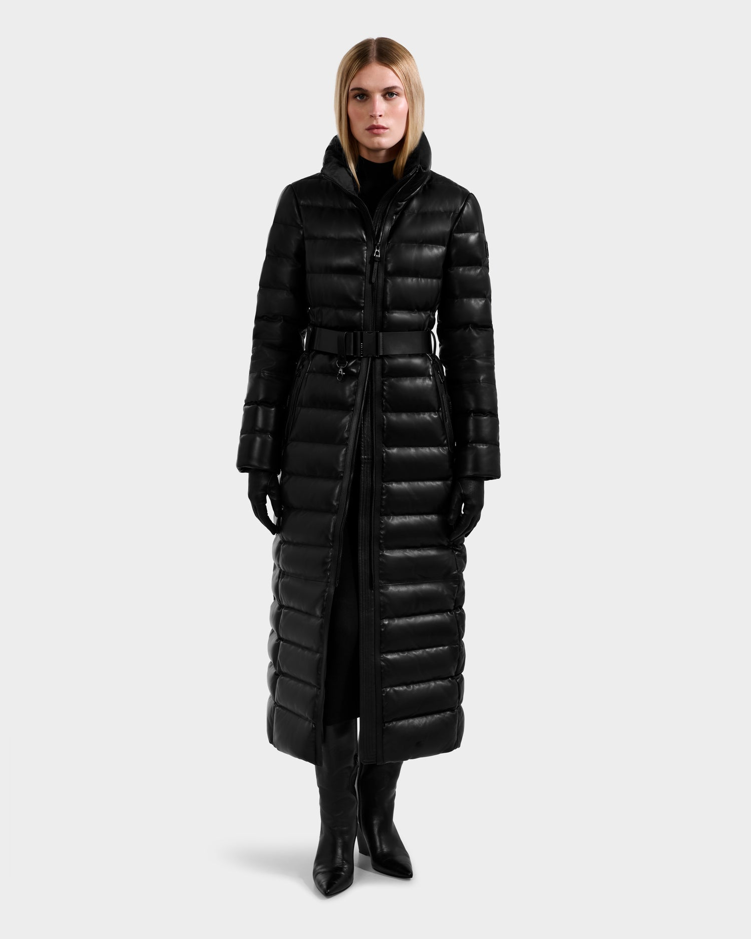 A person stands facing forward in the Rudsak PELIN LX - 8125692 Black women’s maxi down puffer coat with a belt, paired with black gloves, pants, and heeled boots against a plain light grey background.