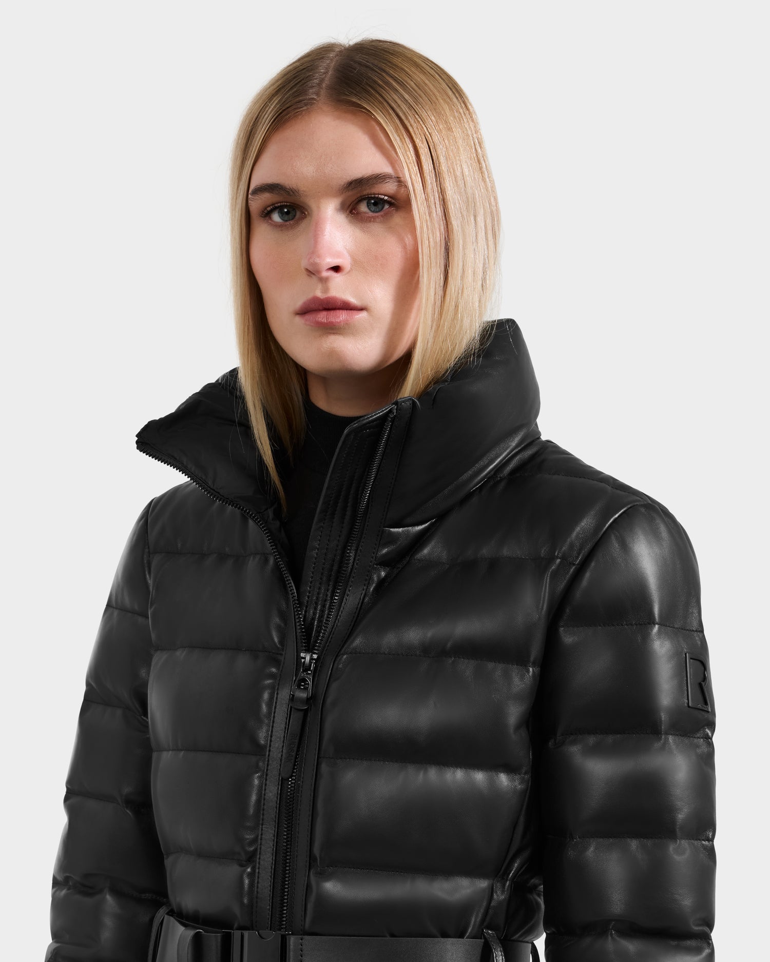 A person with straight blonde hair wears the Rudsak PELIN LX - 8125692 Black leather down puffer jacket with a belted waist and high collar, looking at the camera against a plain light background.