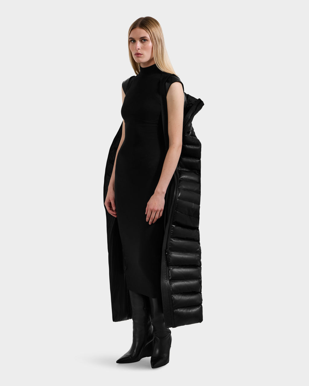 A woman with long blonde hair wears a sleeveless black dress, black heeled boots, and the Rudsak PELIN LX - 8125692 Black maxi coat draped over her shoulders, standing against a plain light gray background.