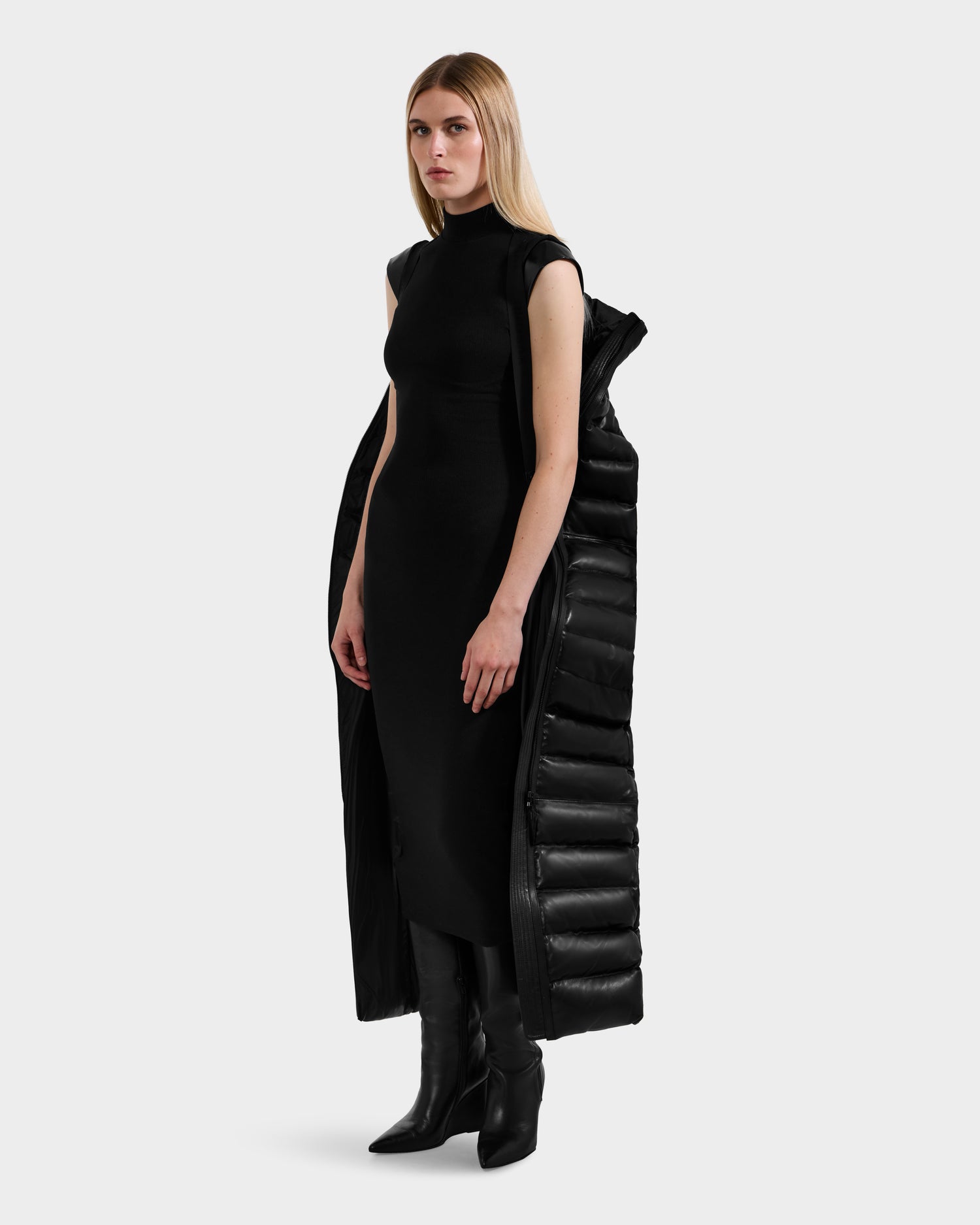 A woman with long blonde hair wears a sleeveless black dress, black heeled boots, and the Rudsak PELIN LX - 8125692 Black maxi coat draped over her shoulders, standing against a plain light gray background.