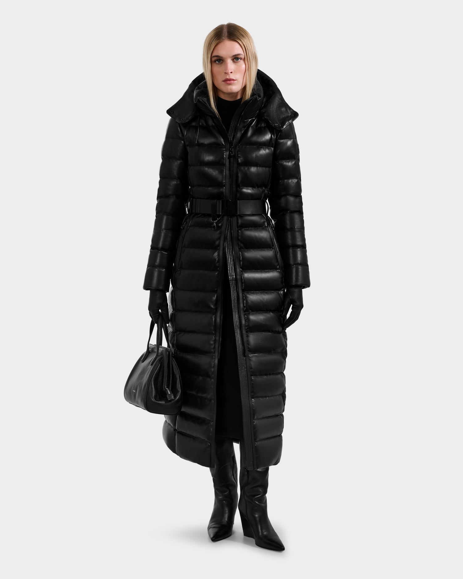 A woman wearing the Rudsak PELIN LX - 8125692 Black long leather down puffer, with black gloves and heeled boots, holds a black handbag against a light gray background, looking directly at the camera.