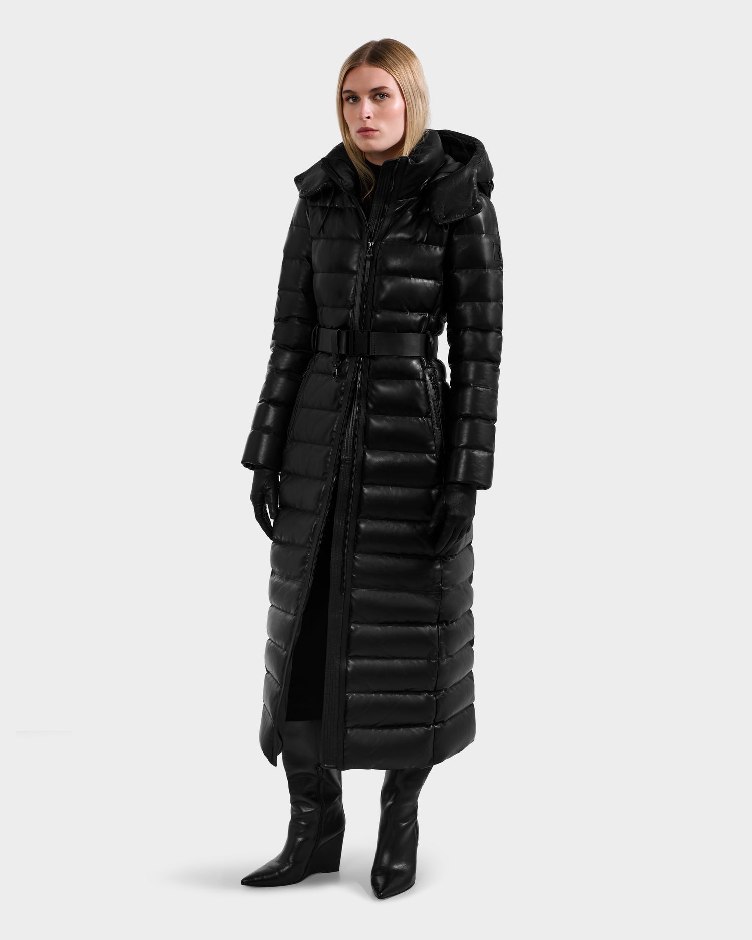 A woman with straight, light brown hair wears the Rudsak PELIN LX - 8125692 Black leather down puffer with a hood, paired with black gloves and heeled boots, posing against a plain light gray background.