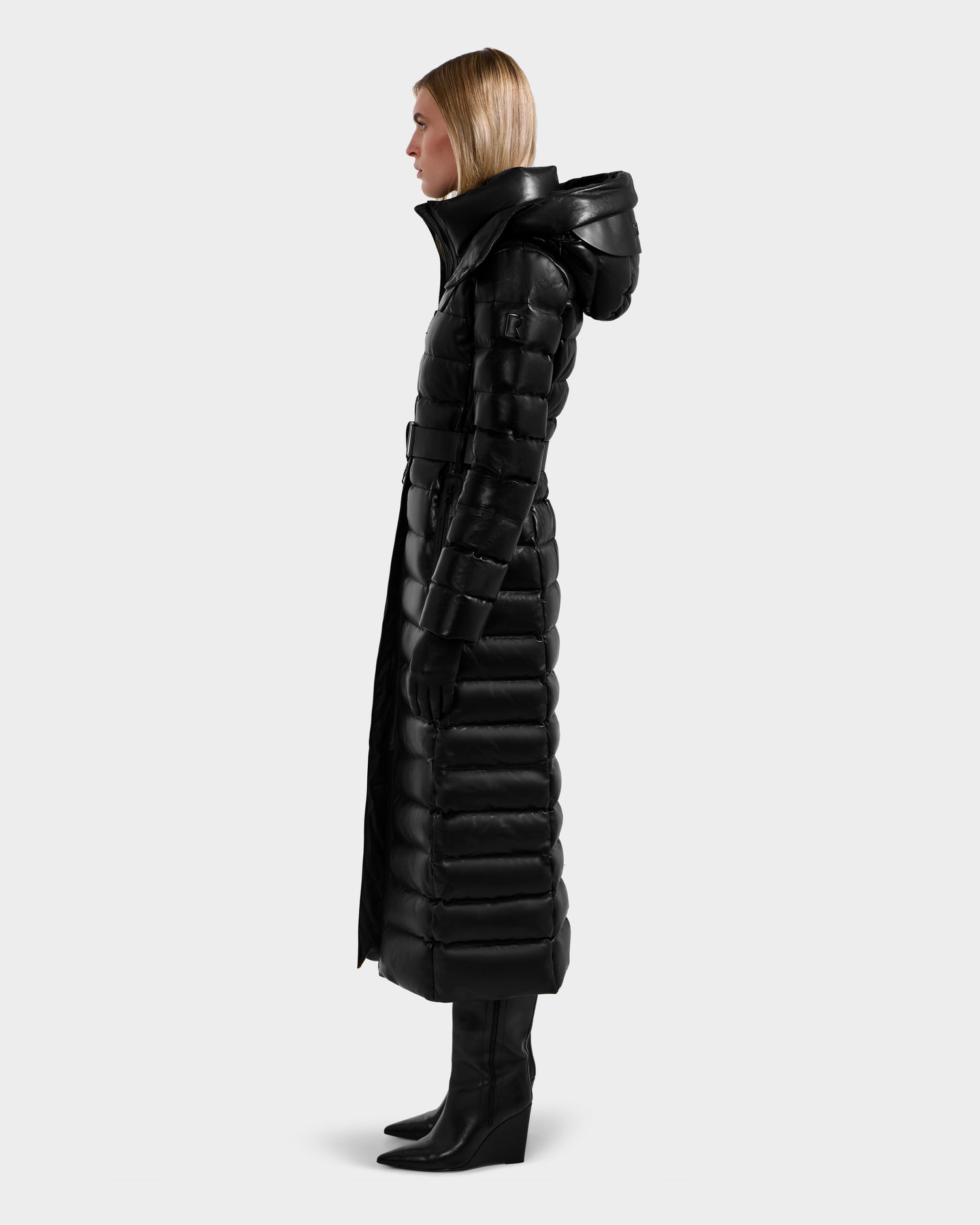 A person in profile wears the Rudsak PELIN LX - 8125692 Black leather down puffer women’s maxi coat with a hood, paired with black gloves and knee-high boots, standing against a plain light gray background.