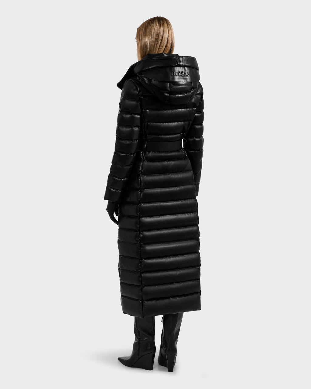 A woman faces away, wearing the Rudsak PELIN LX - 8125692 Black maxi coat—a long black leather down puffer with a hood—paired with black gloves and knee-high heeled boots against a plain light gray background.