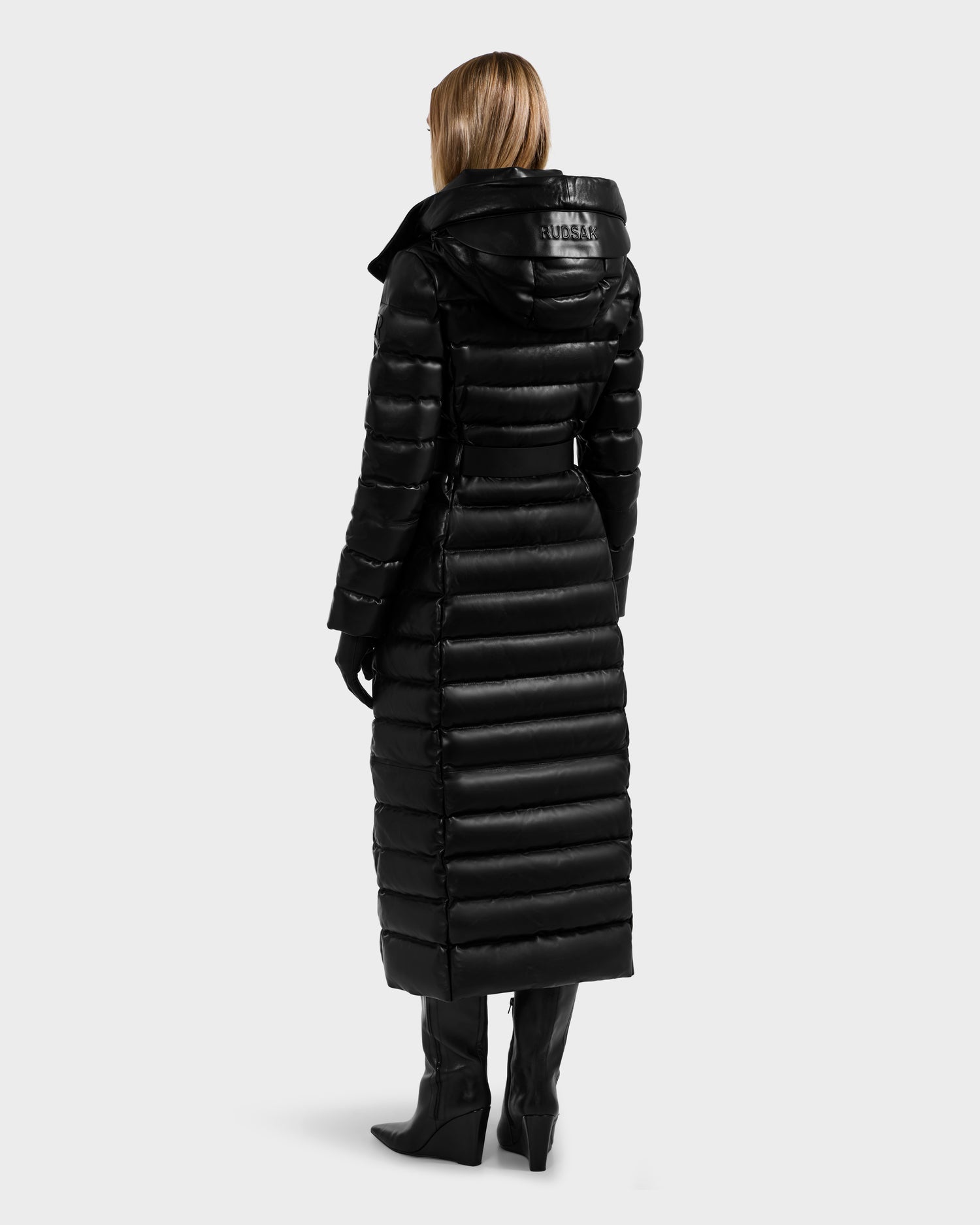 A woman faces away, wearing the Rudsak PELIN LX - 8125692 Black maxi coat—a long black leather down puffer with a hood—paired with black gloves and knee-high heeled boots against a plain light gray background.