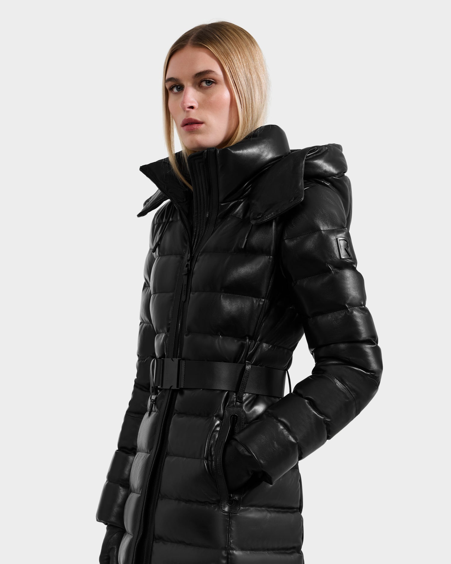 A woman with straight blonde hair models the Rudsak PELIN LX - 8125692 Black women’s maxi coat—a shiny black leather down puffer with a high collar, belted waist, and hood—against a plain light gray background.