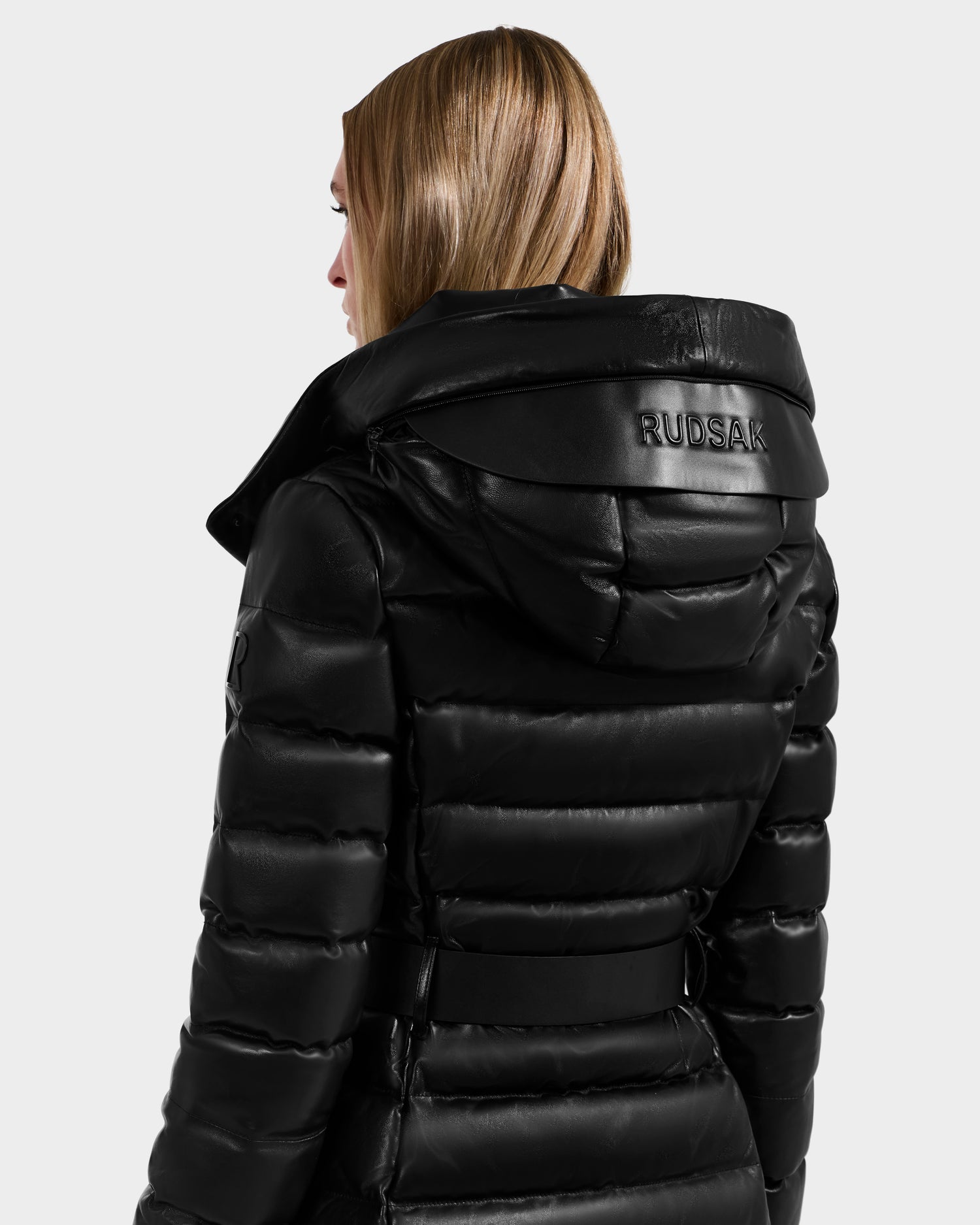 A person with straight, light brown hair wears the Rudsak PELIN LX - 8125692 Black women’s maxi down puffer—a belted, leather coat with a large hood featuring the brand name embossed on the back. The individual is photographed from behind.