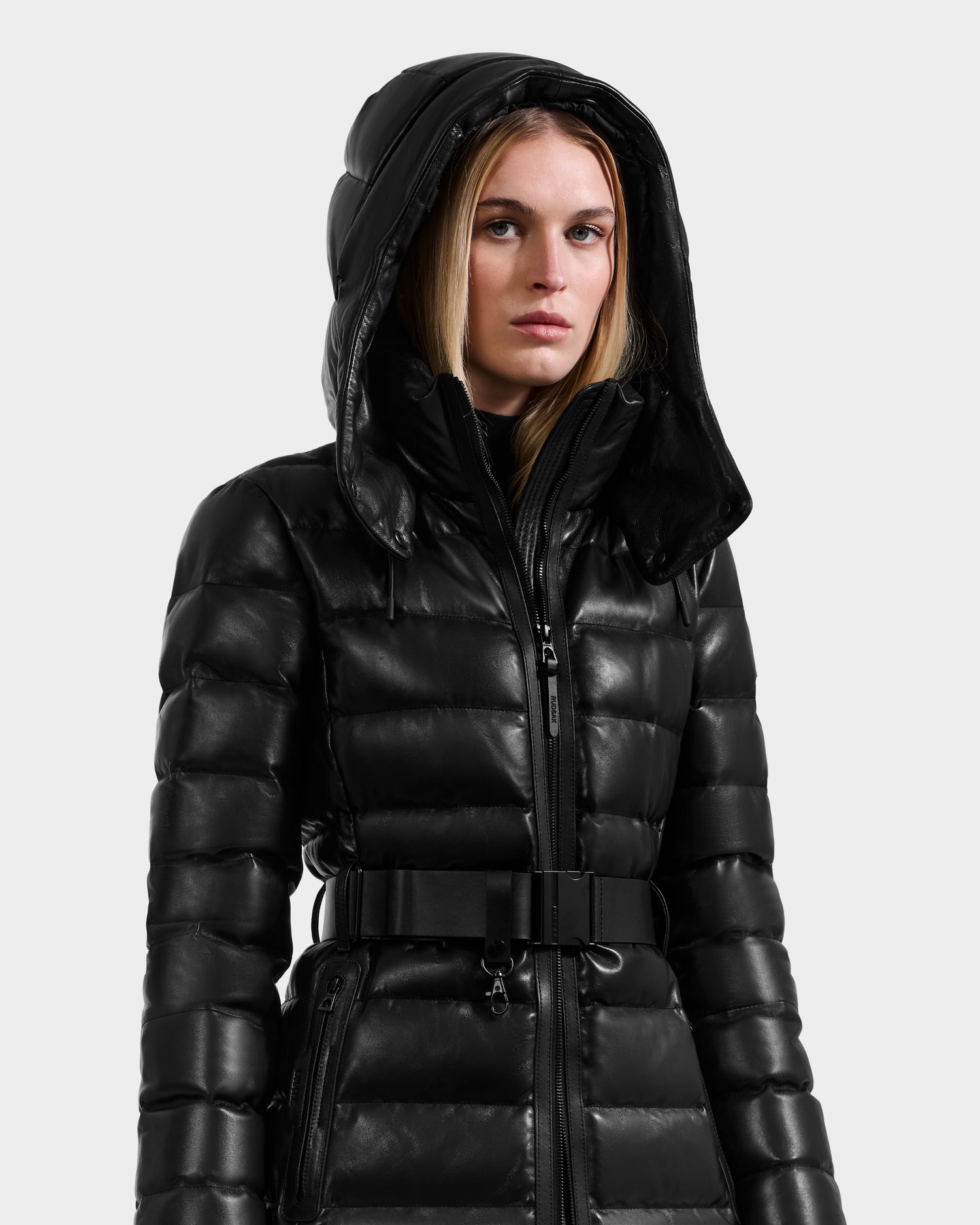 A woman models the Rudsak PELIN LX - 8125692 Black leather down puffer jacket with hood, zippered front, and waist belt against a plain light gray background.