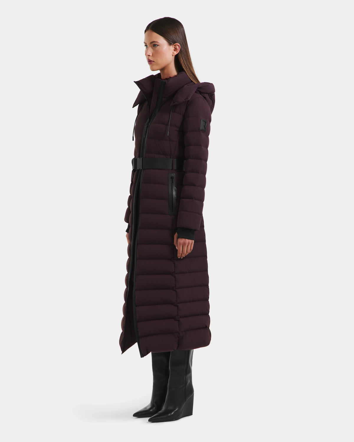 A woman stands in profile wearing the Rudsak PELIN L - 8124815 Bordeaux Mocha, a long down puffer with belted waist and hood, paired with black knee-high boots against a plain white background.