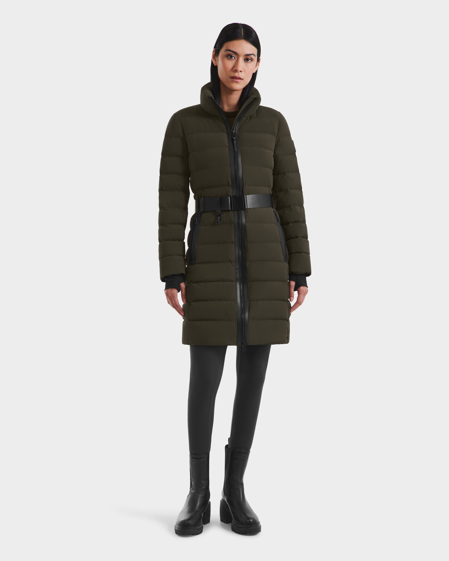 A woman with straight dark hair and a neutral expression faces forward in a Rudsak PELIN M - 8124814 Dark Olive down puffer with a belt, paired with black leggings and ankle boots. The background is plain white.