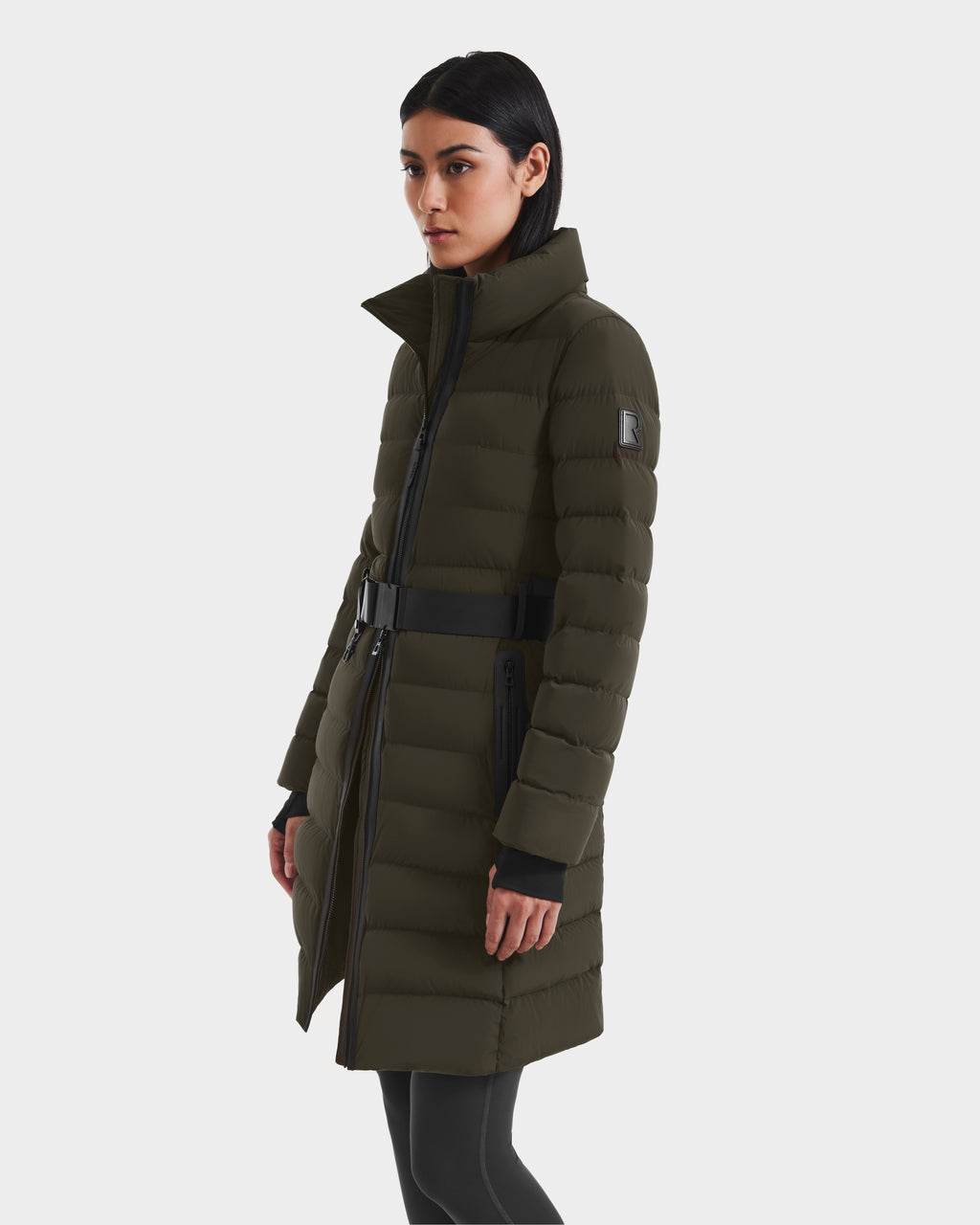 A woman wearing the Rudsak PELIN M - 8124814 Dark Olive RDS-certified down puffer with a belted waist and black zipper details pairs it with black leggings, standing against a plain white background.