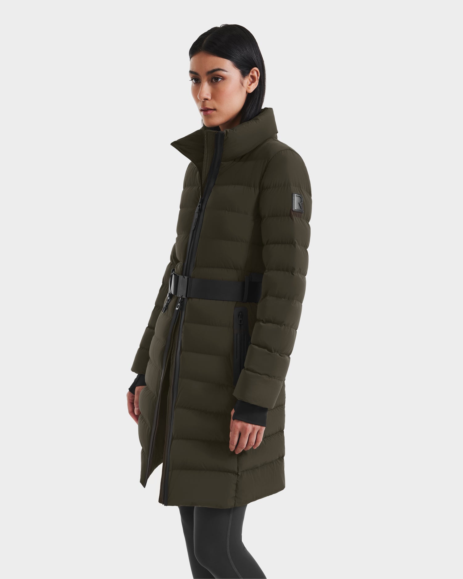 A woman wearing the Rudsak PELIN M - 8124814 Dark Olive RDS-certified down puffer with a belted waist and black zipper details pairs it with black leggings, standing against a plain white background.
