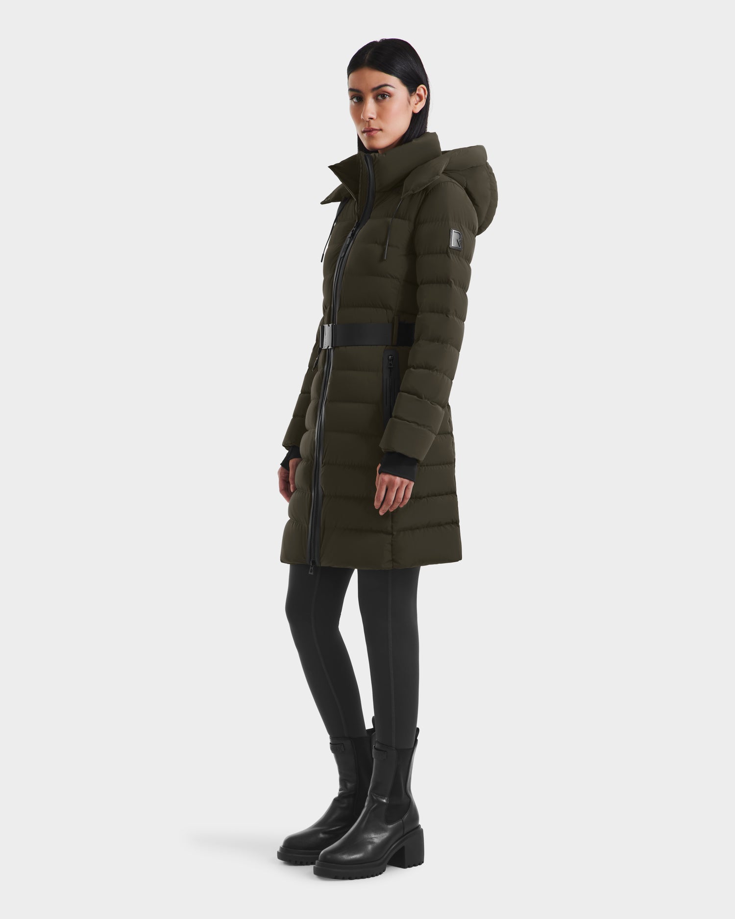 A woman in the Rudsak PELIN M - 8124814 Dark Olive down puffer with a hood, black leggings, and black ankle boots stands against a plain white background.