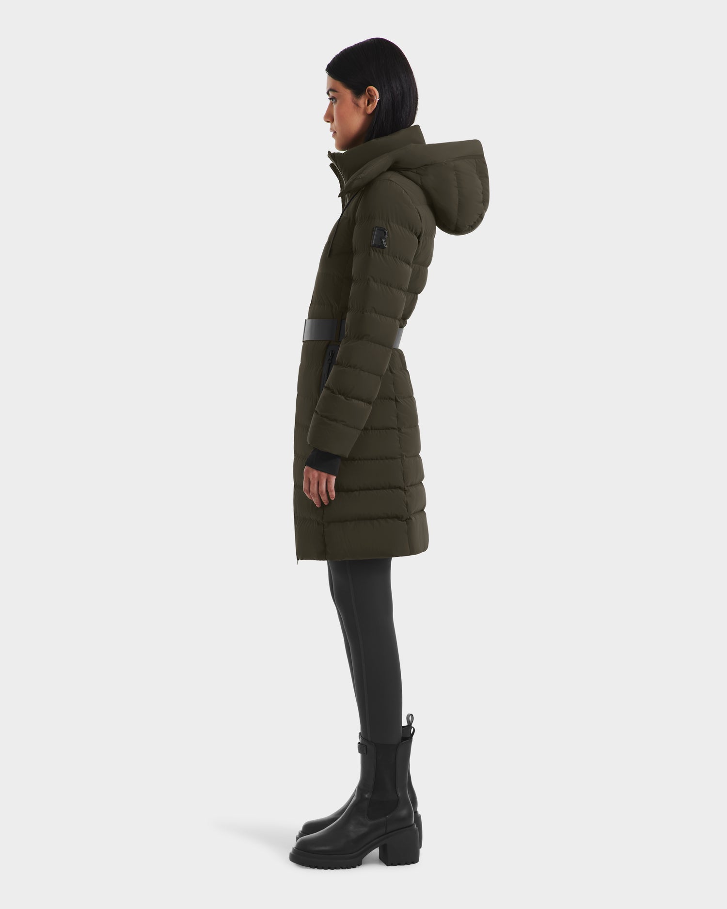 A woman in profile wears the Rudsak PELIN M - 8124814 Dark Olive womens down puffer coat with a hood, black leggings, and black boots, standing against a plain white background.