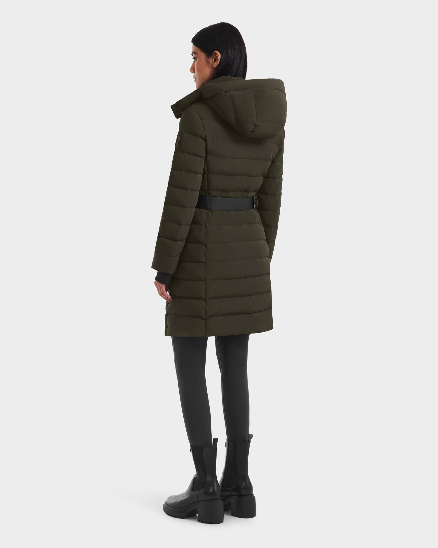 A woman faces away in a Rudsak PELIN M - 8124814 Dark Olive RDS-certified women’s down puffer with a belt, paired with black leggings and chunky boots, set against a plain white background.