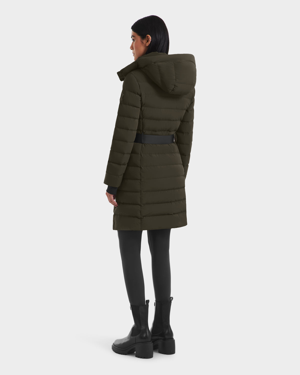 A woman faces away in a Rudsak PELIN M - 8124814 Dark Olive RDS-certified women’s down puffer with a belt, paired with black leggings and chunky boots, set against a plain white background.