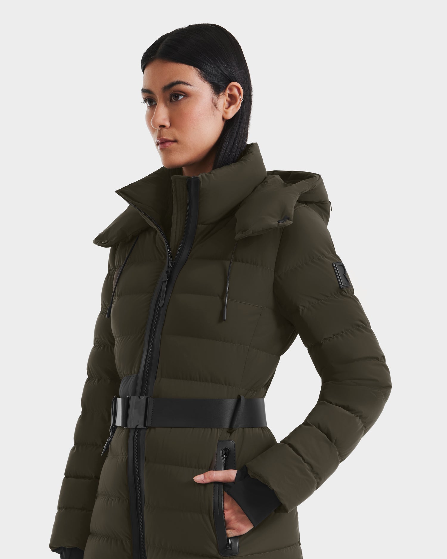 A woman in the Rudsak PELIN M - 8124814 Dark Olive, an RDS-certified women's down puffer coat with a high collar and zippered pockets, stands against a plain white background, looking to the side.