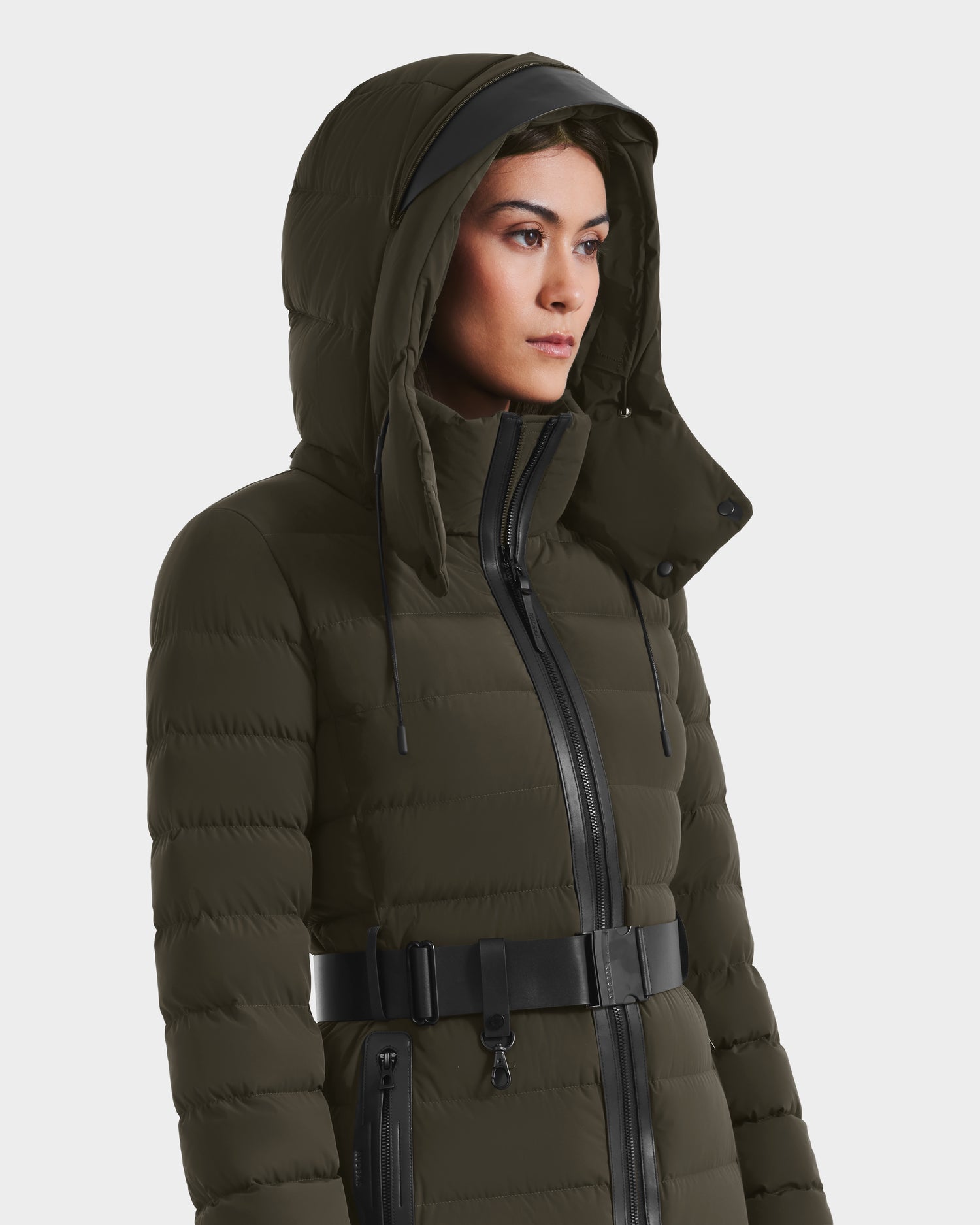 A woman stands facing slightly right against a white background, wearing the Rudsak PELIN M - 8124814 Dark Olive RDS-certified quilted winter jacket with a wide black belt and zippered front.