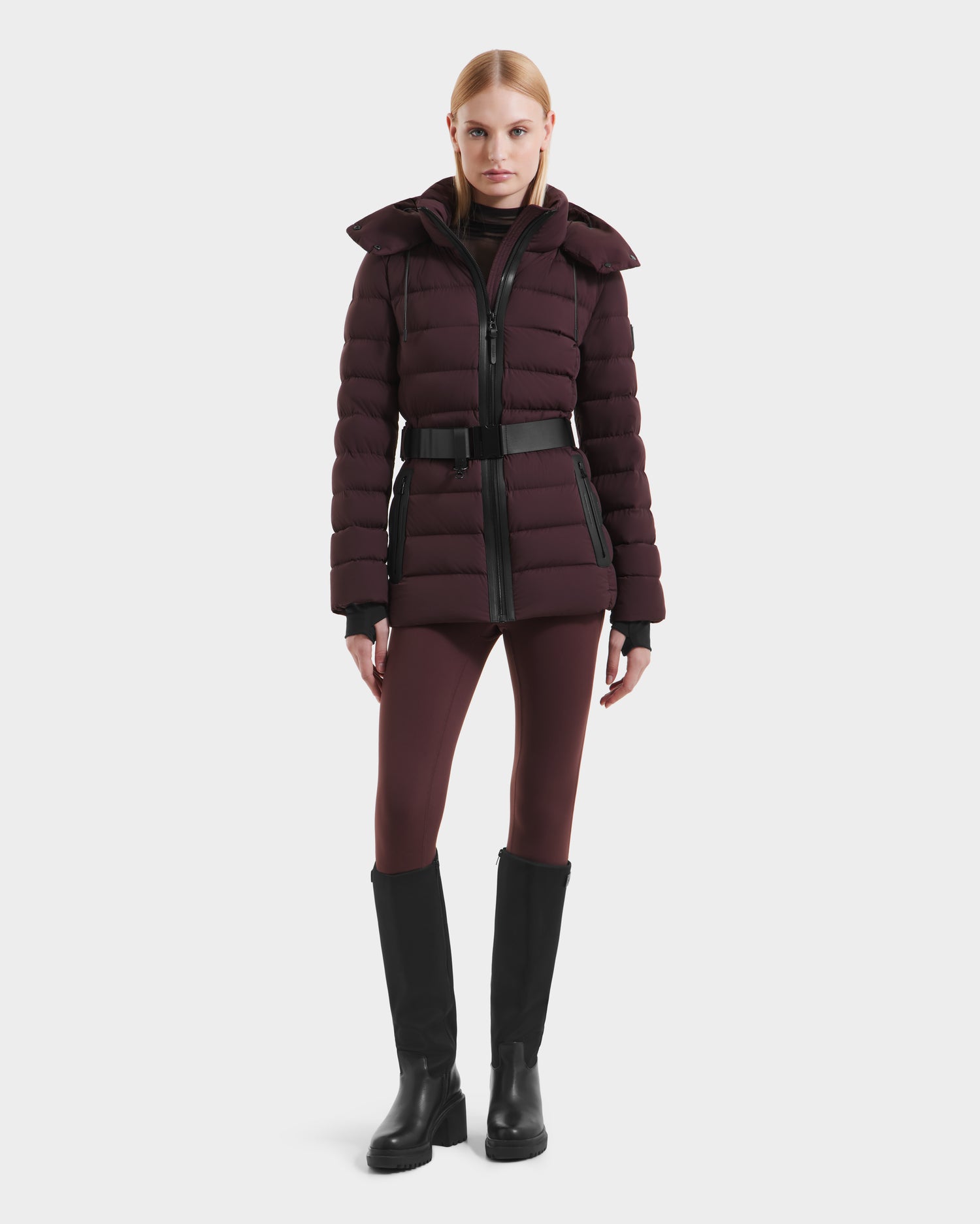 A woman faces forward against a plain white background, wearing the Rudsak PELIN S - 8124813 down puffer in Bordeaux, paired with matching leggings, black boots, and a black turtleneck.