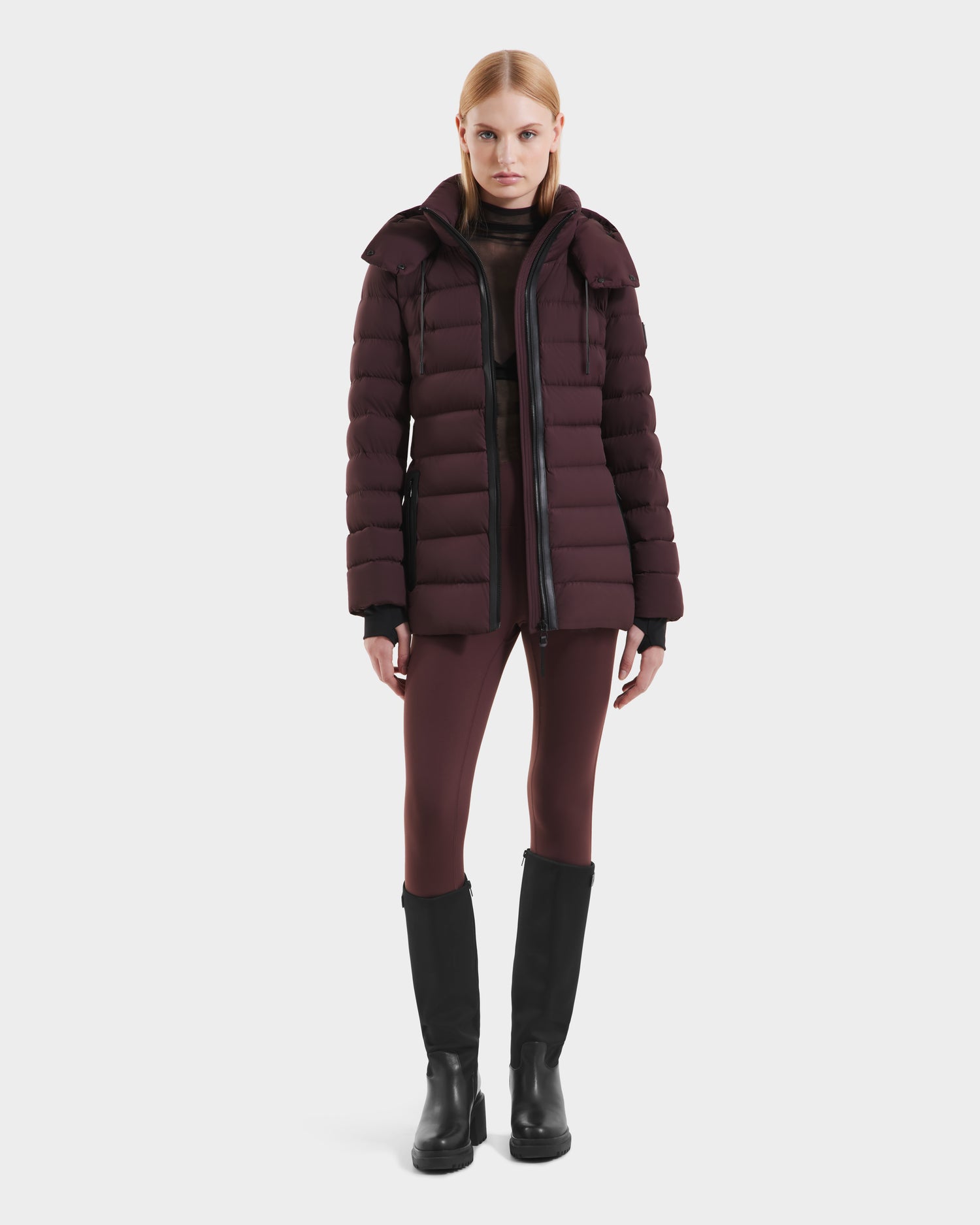A woman with straight, shoulder-length blonde hair stands against a white background wearing the Rudsak PELIN S - 8124813 Bordeaux women’s down puffer jacket, matching leggings, a black sheer top, and black knee-high boots.