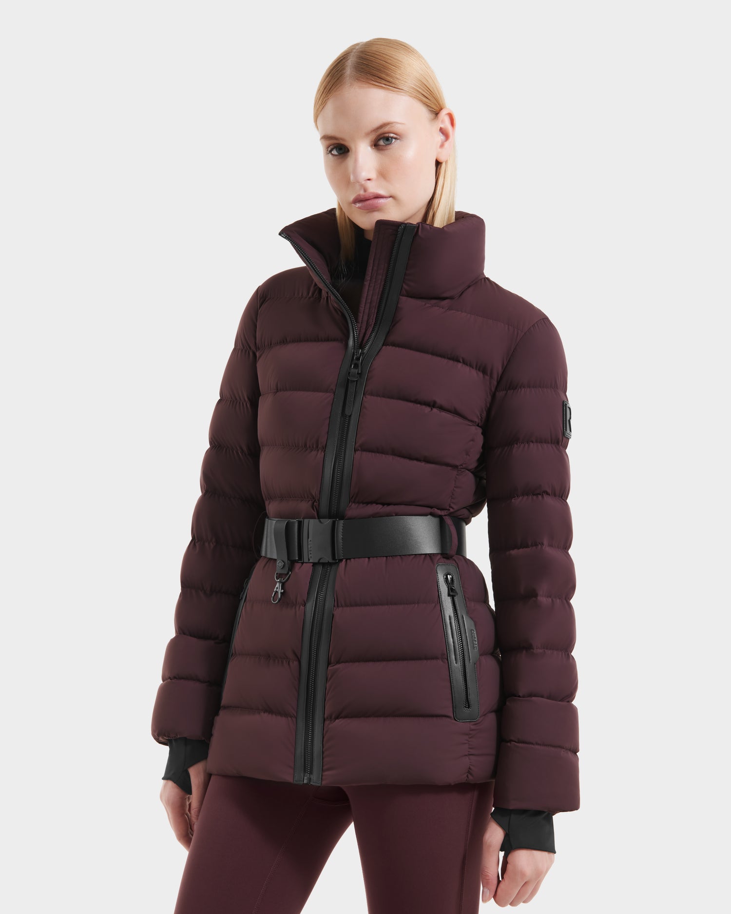 A woman stands against a white background wearing the Rudsak PELIN S - 8124813 Bordeaux down puffer with a high collar, black zipper, and matching belt, her hands at her sides.