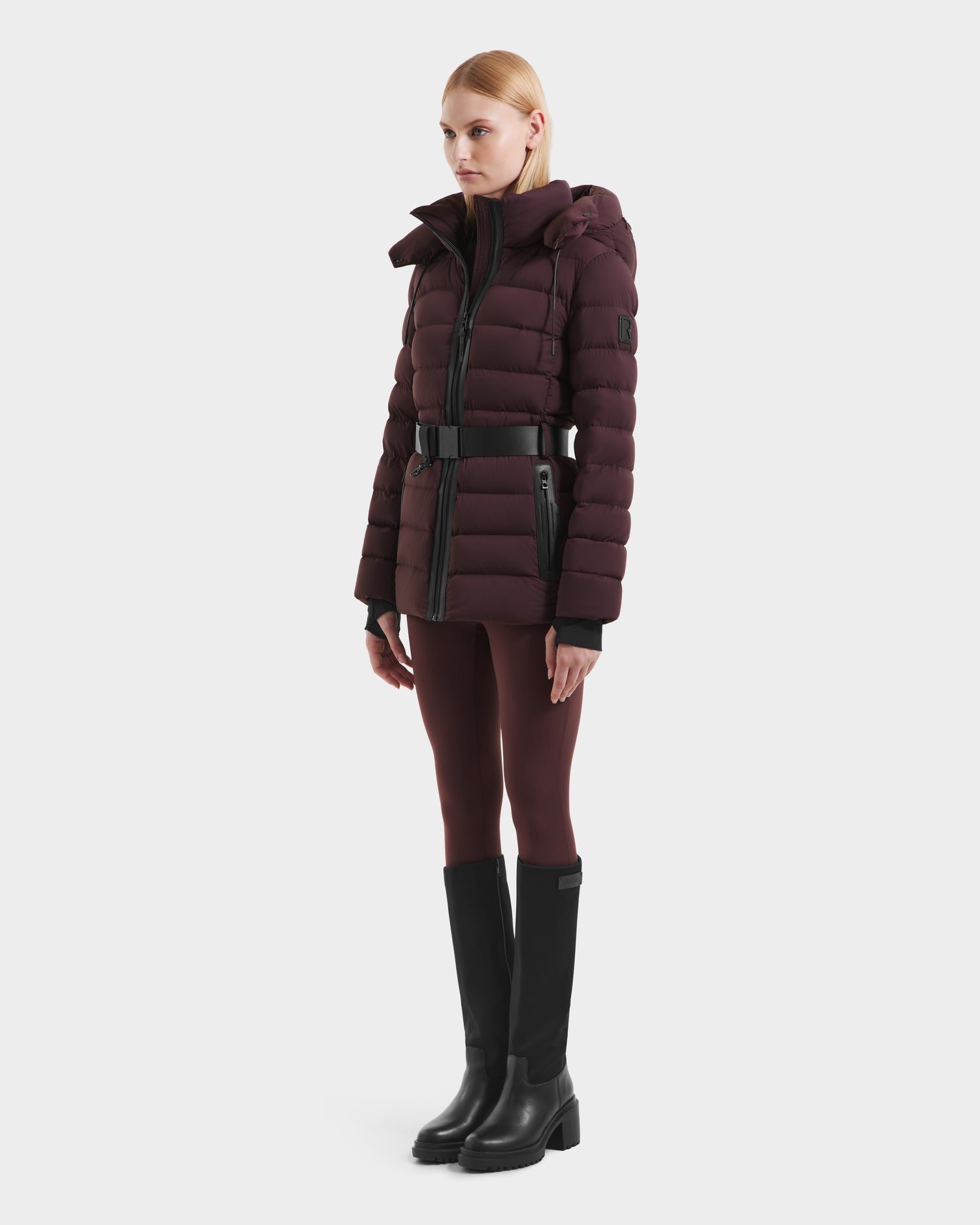 A person stands slightly turned, wearing the Rudsak PELIN S - 8124813 Bordeaux womens down puffer jacket with a hood, paired with matching leggings and black knee-high boots, set against a plain white background.