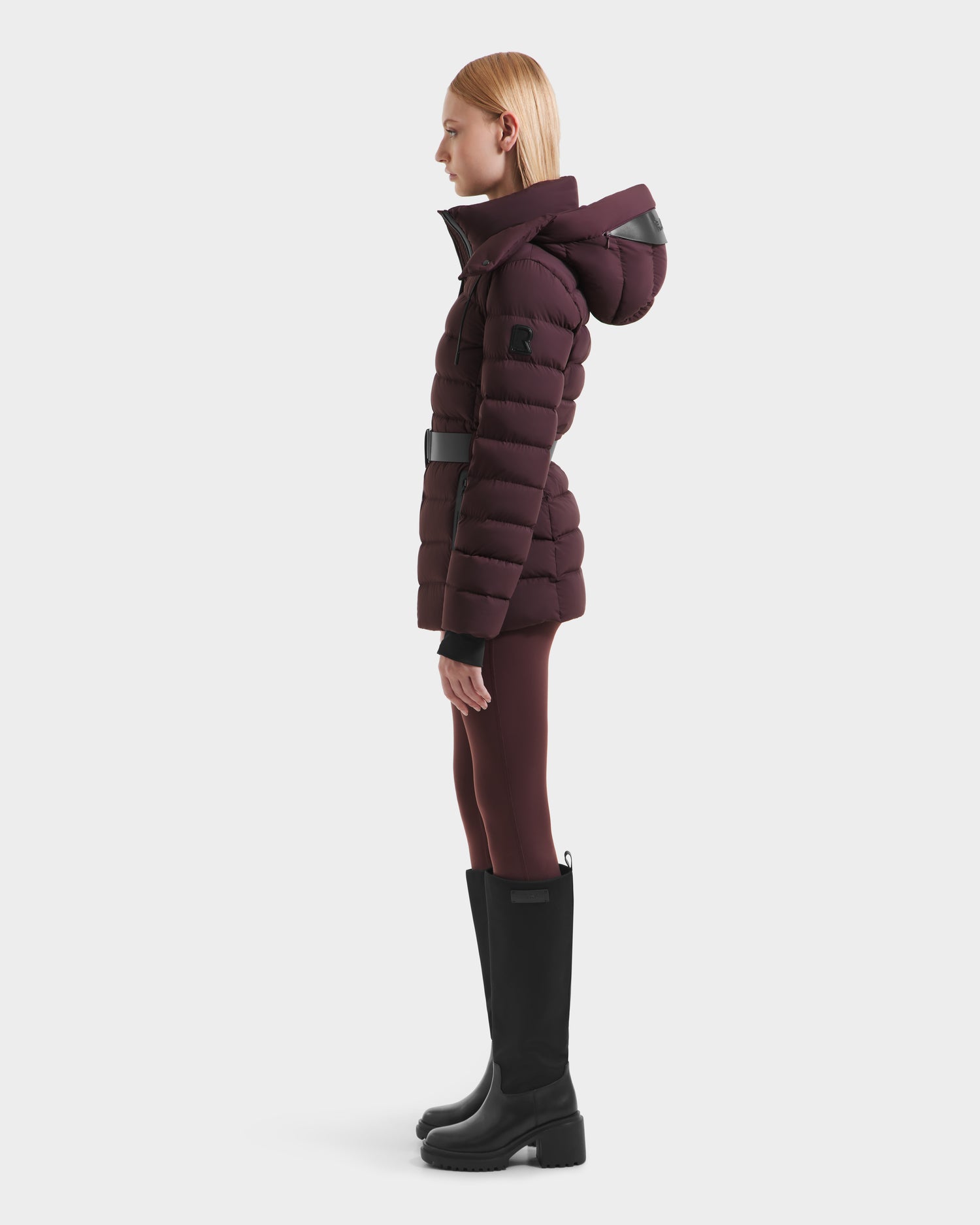 A woman stands in profile against a white background, wearing the Rudsak PELIN S - 8124813 Bordeaux down puffer jacket with a hood, coordinating leggings, and tall black boots.