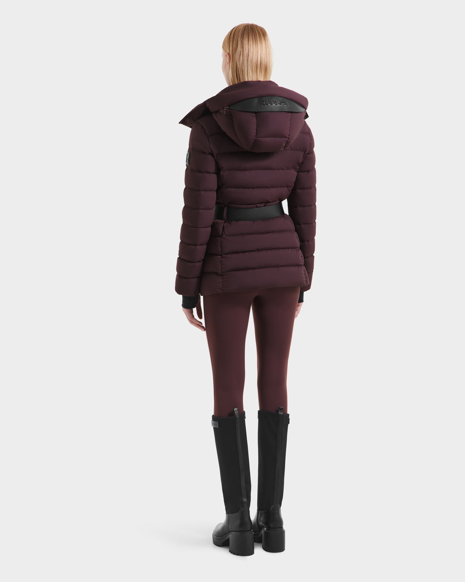 A person with straight blonde hair is seen from behind wearing the Rudsak PELIN S - 8124813 Bordeaux down puffer jacket with matching leggings and tall black boots, set against a plain white background.