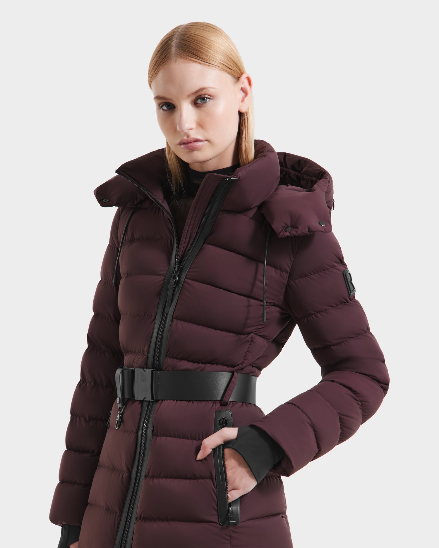 A woman stands confidently against a white background, wearing the Rudsak PELIN S - 8124813 Bordeaux down puffer with a high collar, hood, and black belt.
