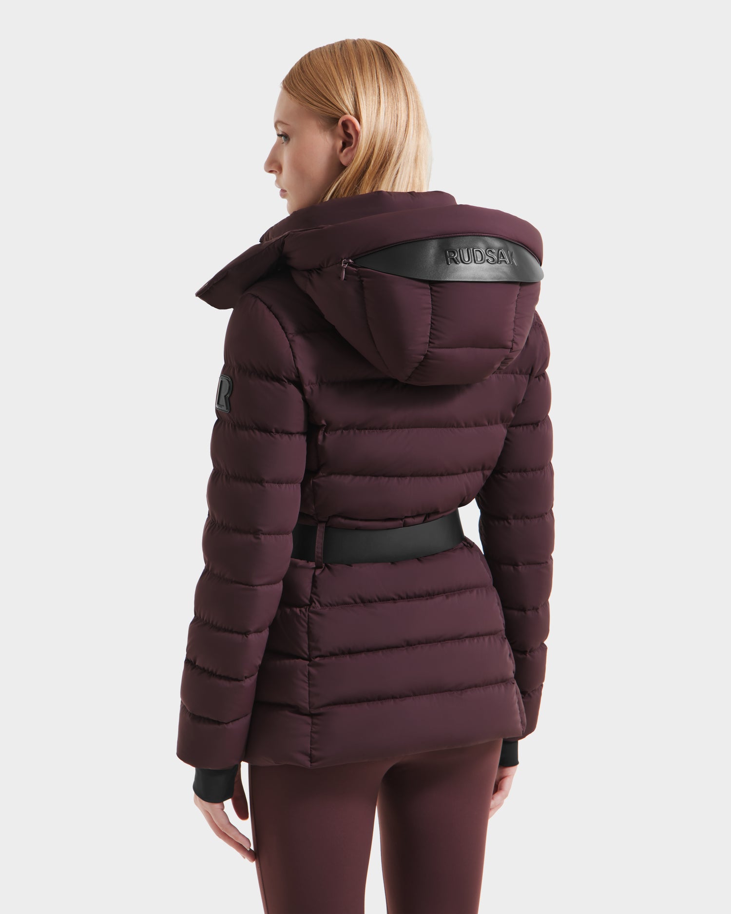 A person with straight, light brown hair wears the Rudsak PELIN S - 8124813 Bordeaux women’s down puffer jacket, featuring a hood, black belt, and arm patch. Shown from the back as the person faces away. RDS-certified down.