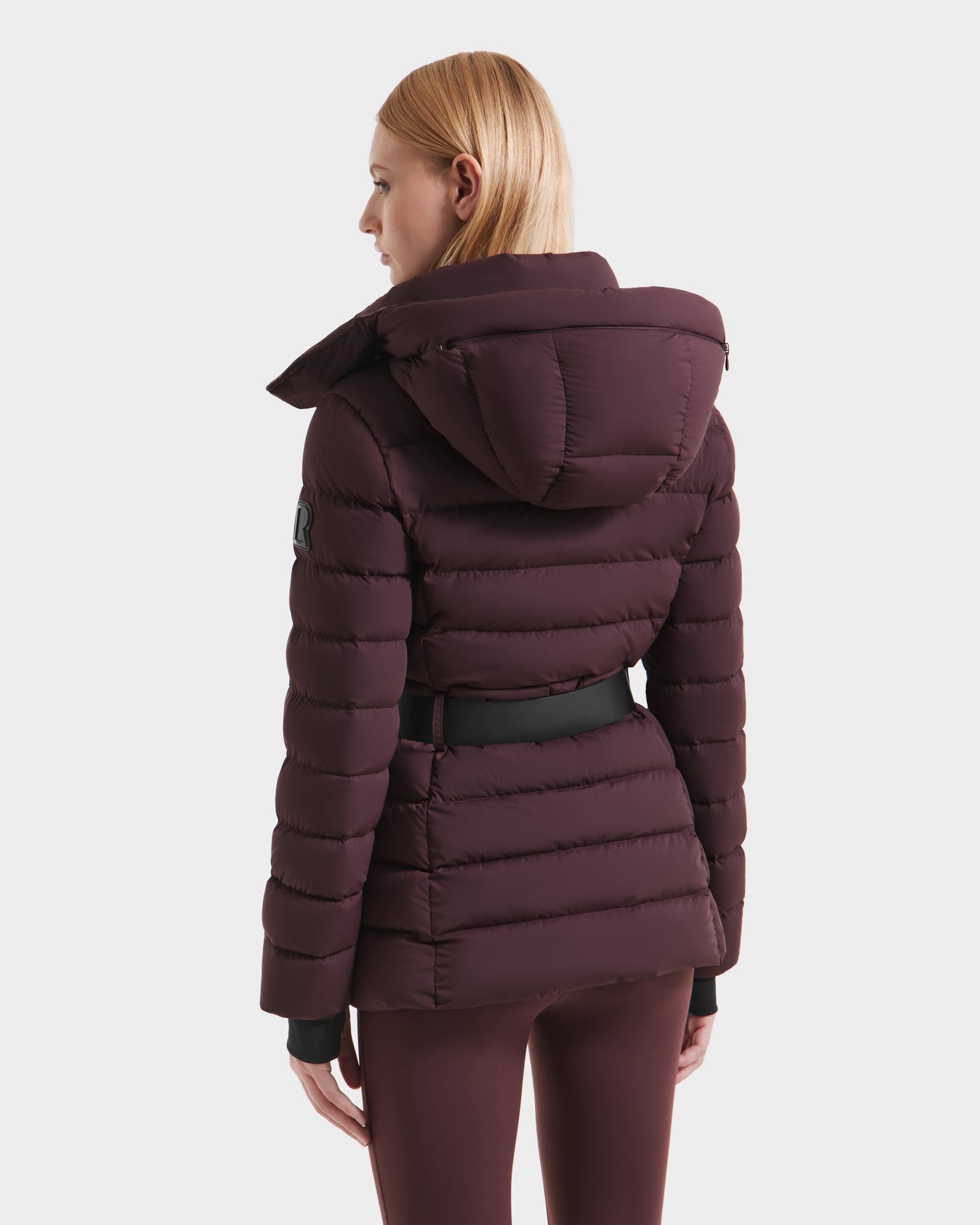 A woman with straight, shoulder-length blonde hair is seen from the back wearing the Rudsak PELIN S - 8124813 Bordeaux womens down puffer jacket with hood and black belt, paired with matching bordeaux pants.