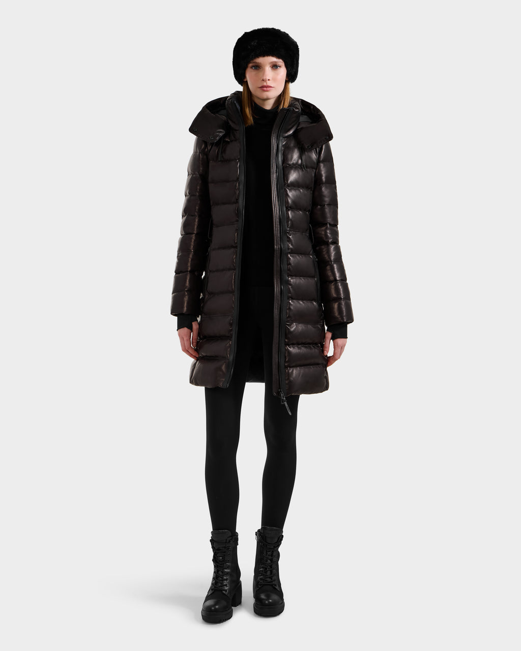 A woman faces forward in a shiny black Rudsak PELIN X - 8125575 down puffer coat, paired with black leggings, lace-up boots, and a black fur hat against a plain light background.