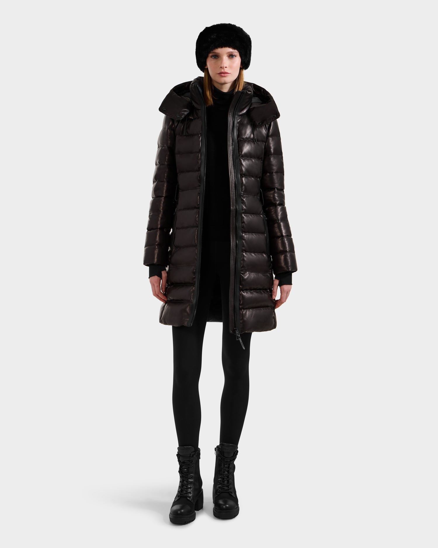 A woman faces forward in a shiny black Rudsak PELIN X - 8125575 down puffer coat, paired with black leggings, lace-up boots, and a black fur hat against a plain light background.
