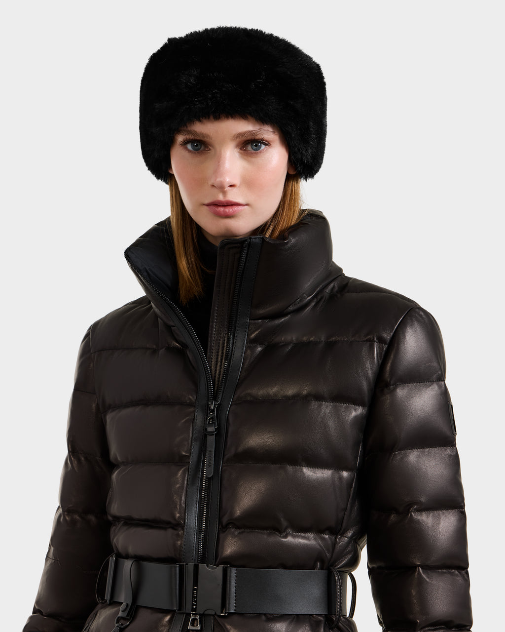 A woman in a shiny black Rudsak PELIN X - 8125575 leather down puffer with a high collar and wide belt, paired with a black faux fur hat, stands against a plain light background.