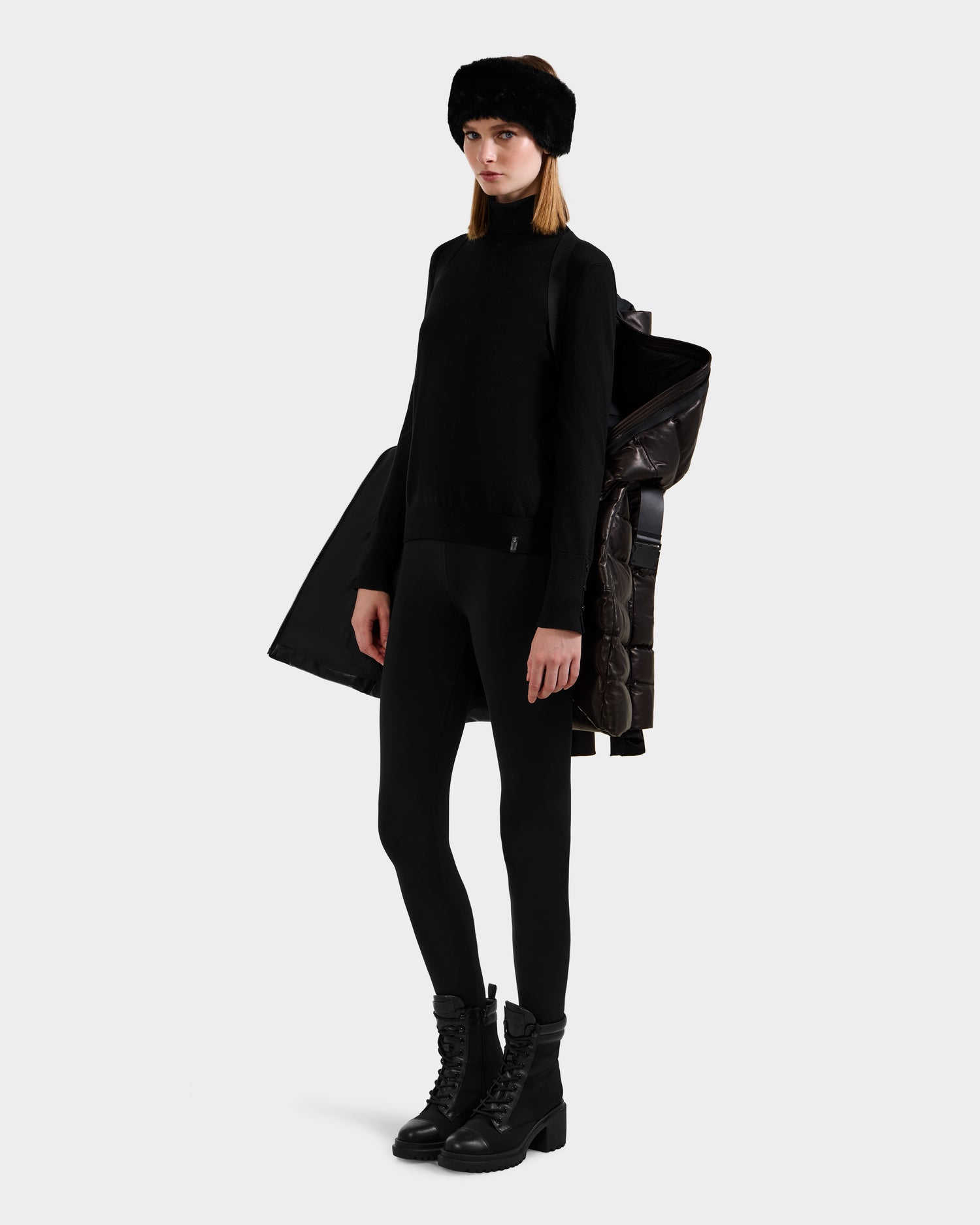 A woman in an all-black outfit with a pullover, leggings, lace-up boots, and a fur headband poses holding the Rudsak PELIN X - 8125575 Black leather down puffer women's winter coat draped over her shoulder against a plain light background.