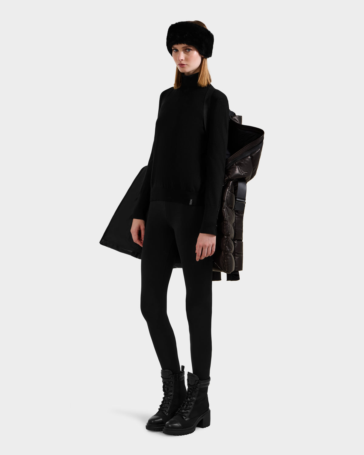 A woman in an all-black outfit with a pullover, leggings, lace-up boots, and a fur headband poses holding the Rudsak PELIN X - 8125575 Black leather down puffer women's winter coat draped over her shoulder against a plain light background.