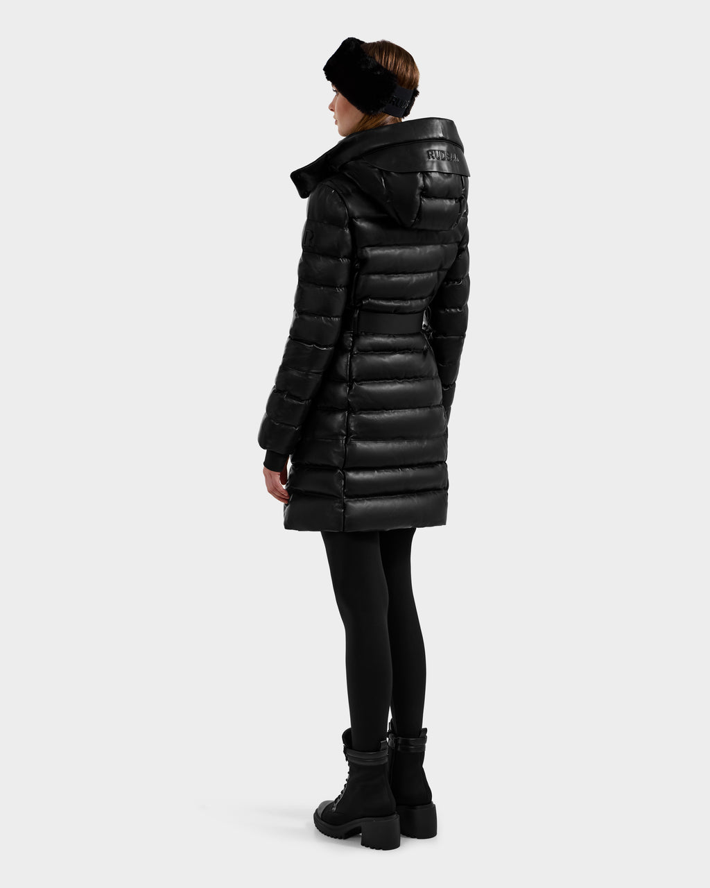 A woman wearing the Rudsak PELIN X - 8125575 Black coat, paired with black leggings, boots, and headband, stands facing away to showcase the back of her quilted down puffer against a plain light background.