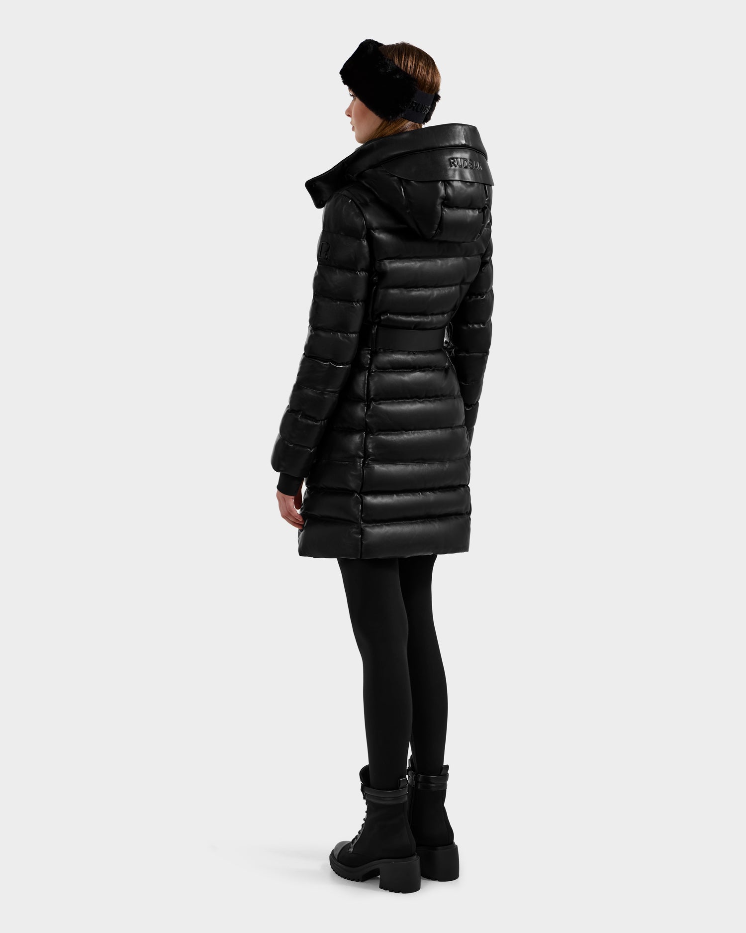 A woman wearing the Rudsak PELIN X - 8125575 Black coat, paired with black leggings, boots, and headband, stands facing away to showcase the back of her quilted down puffer against a plain light background.