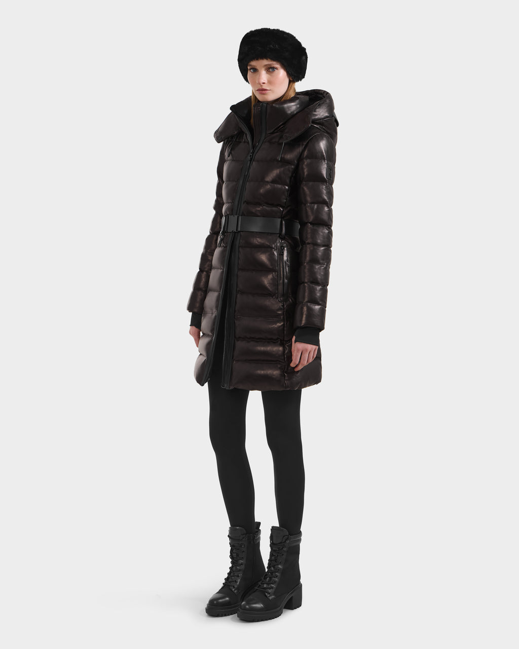 A person wearing the Rudsak PELIN X - 8125575 Black winter coat with a belt, black leggings, lace-up boots, and a black fur hat stands against a plain white background.