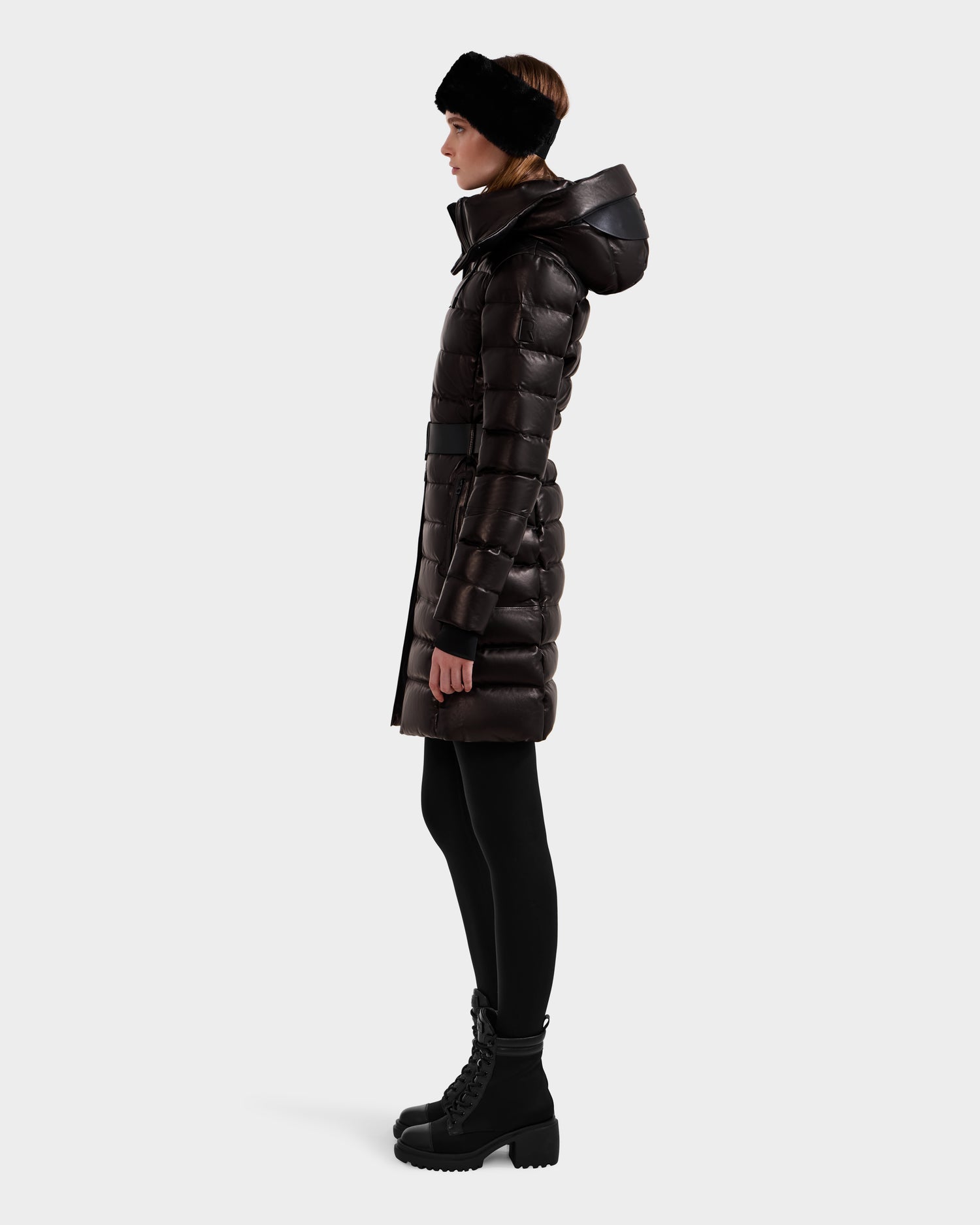 A woman in profile wears the Rudsak PELIN X - 8125575 Black leather down puffer women's winter coat with a hood, paired with black leggings, black lace-up boots, and a black headband against a light background.
