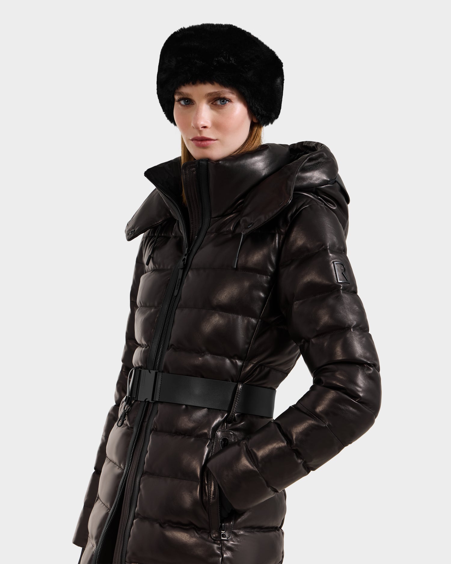 A woman wearing the Rudsak PELIN X - 8125575 Black leather down puffer with a belted waist and a black faux fur hat stands against a plain light background.